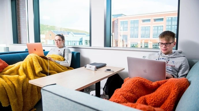 Two young people sitting on sofas with laptops, covered in warm blankets, in a bright room with large windows and a wooden table between them.