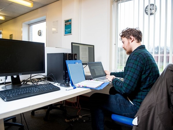 Man with curly hair and beard working on a laptop in an office, sitting at a desk with two desktop monitors and an open blue folder containing papers.