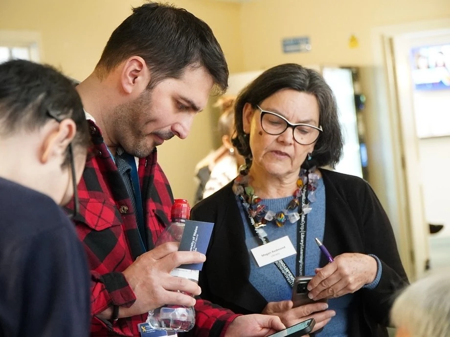A woman with glasses and a colorful necklace showing something on her phone to a man in a red and black checkered shirt holding a water bottle and card, with another person partially visible nearby in an indoor setting.
