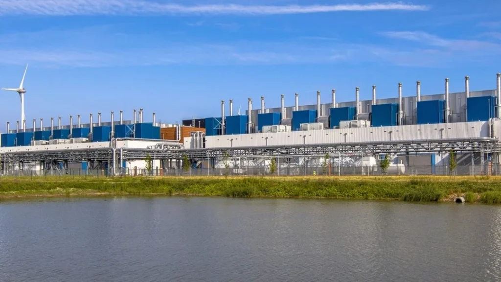 Large industrial facility with multiple exhaust stacks and blue equipment units, situated next to a body of water with a grassy embankment, under a partly cloudy blue sky, with a wind turbine visible on the far left.