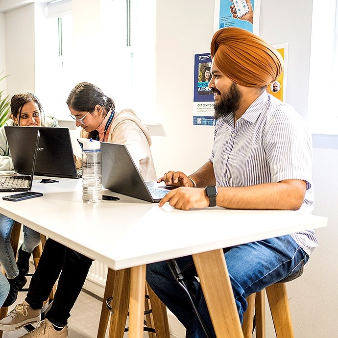 Three people sitting at a white table working on laptops, one person wearing an orange turban and striped shirt smiling, the others focused on their screens, in a bright room with posters on the wall and a clear water bottle on the table.