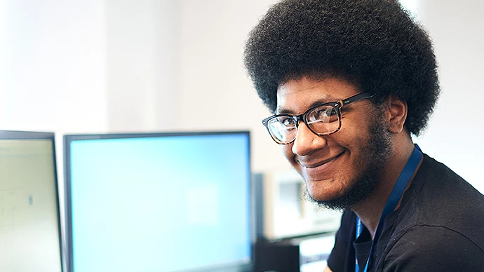 Young man with an afro and glasses smiling while sitting in front of computer monitors in a bright office setting