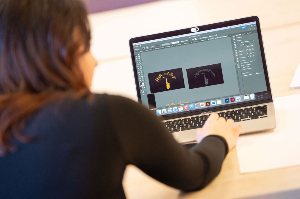 Person working on a MacBook Air with graphic design software open, displaying two similar logos featuring a saxophone and the text 'Rhythm and Blues'.