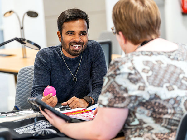 A smiling man holding a pink stress ball while talking to a woman seated across from him in an office setting.