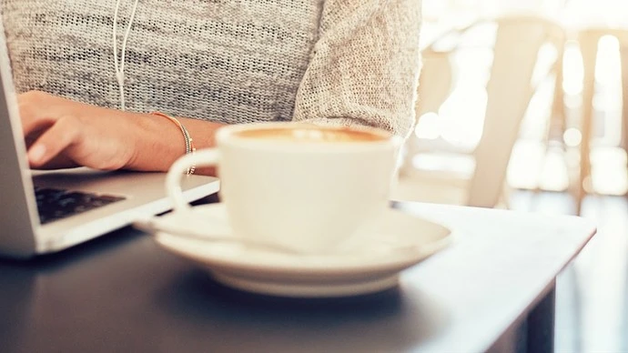 Person typing on a laptop with a white coffee cup and saucer in the foreground on a table in a softly lit indoor setting