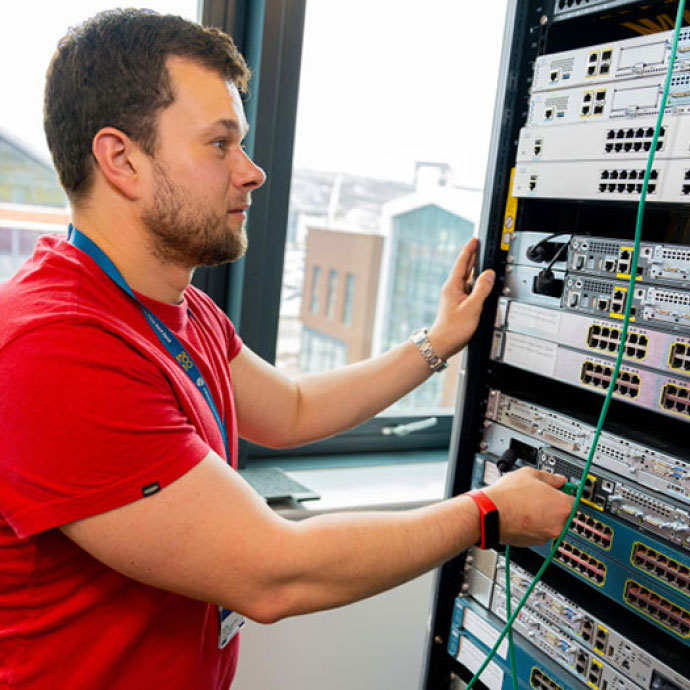 Man in a red shirt working on a network server rack with multiple switches and network devices, adjusting cables inside an office near a window with buildings visible outside.