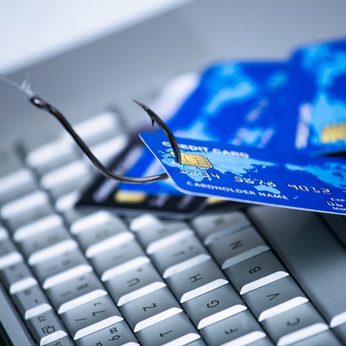Close-up of a fishing hook piercing a blue credit card placed over a computer keyboard, symbolizing credit card phishing or cyber fraud.