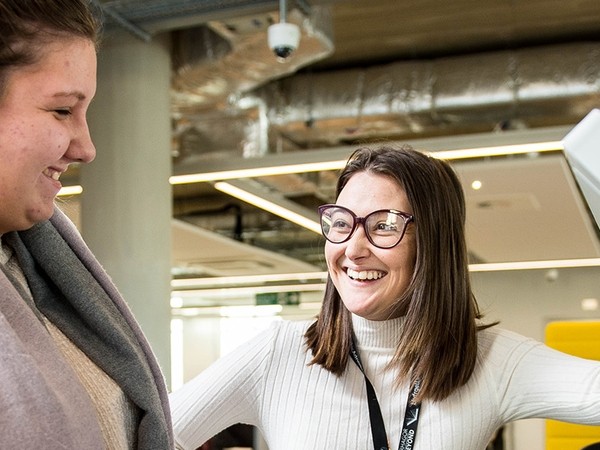 Two women smiling and engaging in conversation indoors, with one wearing glasses and a white sweater and the other in a gray scarf and beige sweater, against a background of modern office ceiling fixtures.