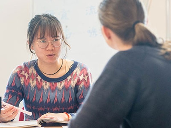 Two people having a conversation across a table, with one person wearing glasses and a patterned sweater speaking while holding a pen and an open book in front of her.