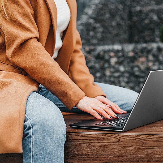 Person wearing a brown coat and blue jeans sitting on a wooden bench, typing on a laptop.