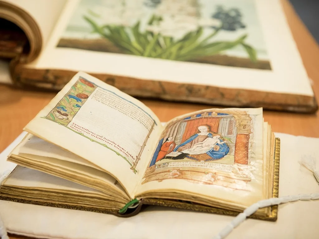 Open illuminated manuscript showing a religious scene of the Virgin Mary holding baby Jesus, with decorative borders and text on the adjacent page, placed on a table with a bead chain and another book in the background.