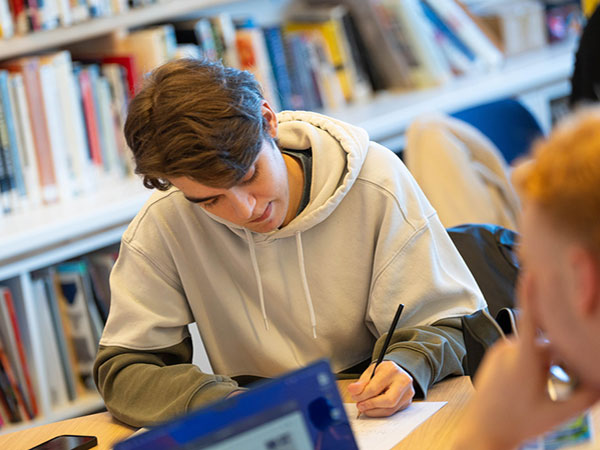 A young person in a beige hoodie writing on paper at a desk in a library with bookshelves in the background and another person blurred in the foreground.