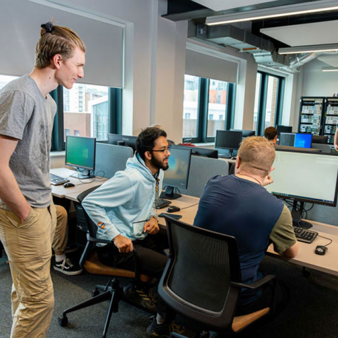 Three young men collaborating in an open-plan office with multiple computer monitors, one standing and two seated, engaging in discussion near a desk with large windows in the background.
