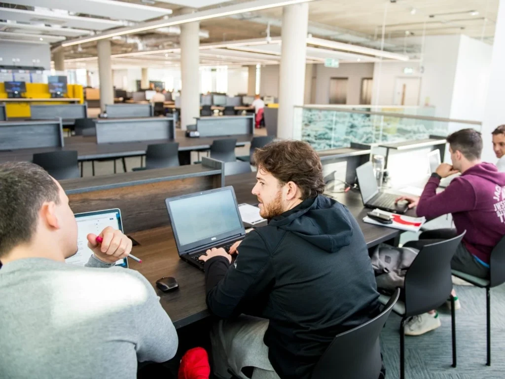 Several young men working on laptops at desks in a modern, spacious open-plan office or study area with multiple desks and chairs spread out in the background.
