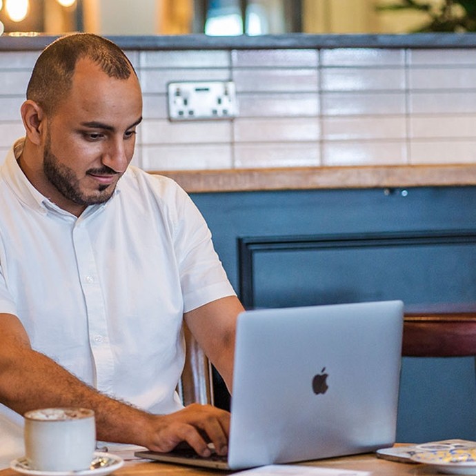 Man with a short beard and short hair wearing a white shirt, sitting at a table using a silver MacBook laptop with a cup of coffee in front of him in a cafe setting.