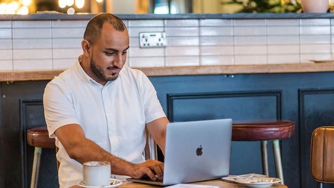 A man in a white shirt sitting at a table in a café, working on a silver MacBook laptop with a cup of coffee nearby.