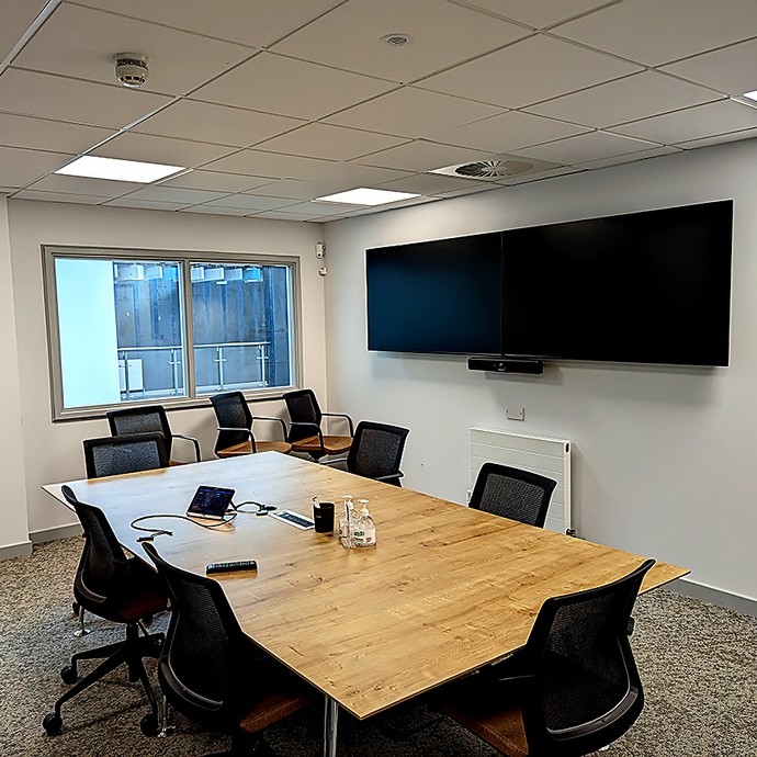 Modern conference room with a large wooden table surrounded by black mesh office chairs, two large flat screen monitors mounted on the wall, a window showing an outdoor area, and various tech accessories on the table.