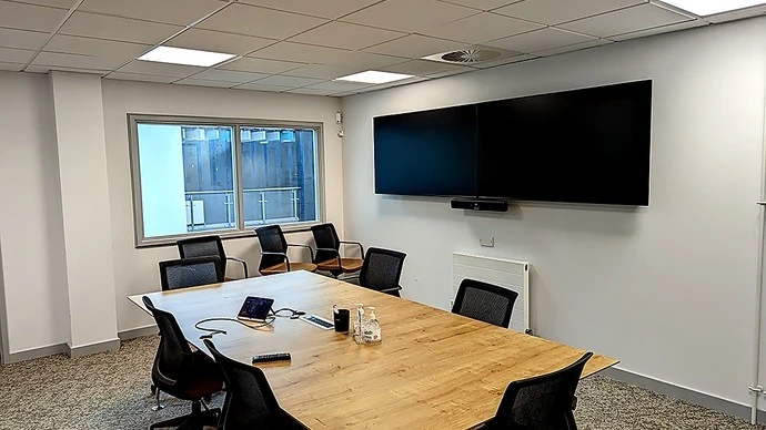 Modern conference room with a long wooden table surrounded by black mesh chairs, a large window on the left wall, and two large flat-screen monitors mounted on the right wall above a white radiator.