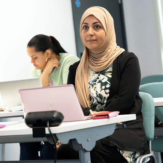 Woman wearing a beige hijab and black cardigan sitting at a desk with a pink HP laptop and a pink notebook, with another woman in the background focused on her work, in a classroom or study setting.