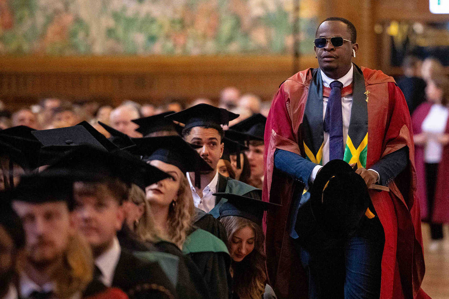 A man wearing sunglasses, a red academic robe with colorful accents, and holding a black hat walks among seated graduates wearing black mortarboards and green graduation gowns in a large hall filled with people.