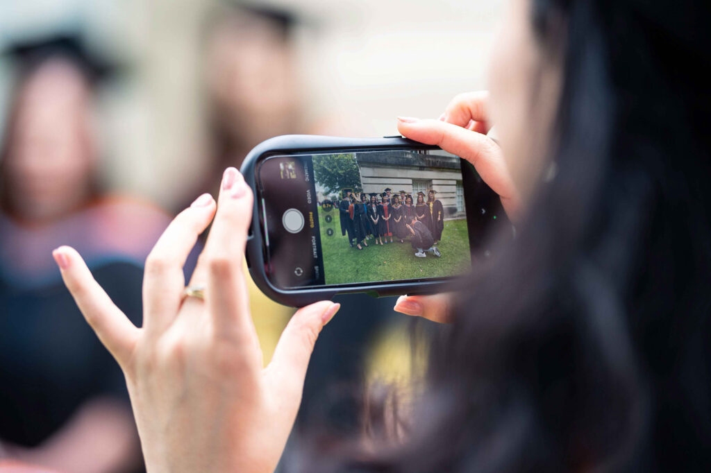 Close-up of a person holding a smartphone horizontally, capturing a photo of a group of graduates in caps and gowns standing on grass in front of a building, with the phone screen showing the graduation photo scene.