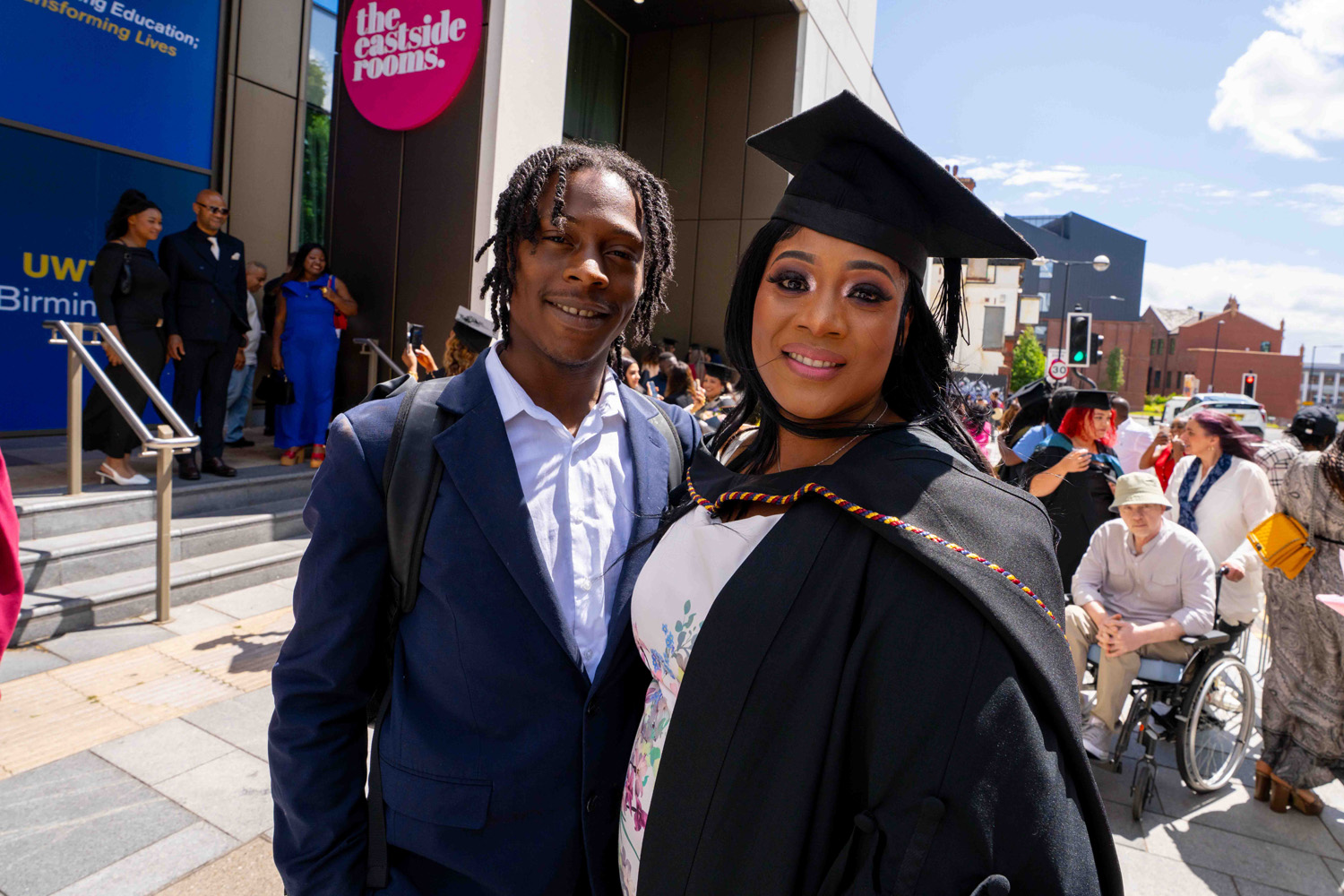 A smiling woman wearing a graduation cap and gown stands next to a man in a dark suit in front of a building with a pink sign that reads 'the eastside rooms,' with a crowd of people behind them including a person in a wheelchair and others celebrating.