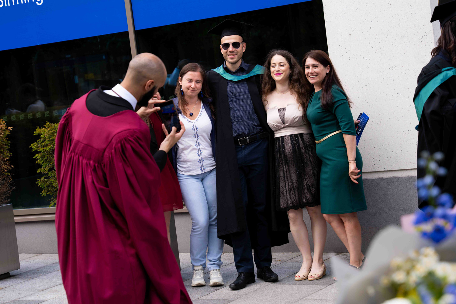 A person in a maroon graduation gown taking a photo with a smartphone of a group of four people standing side by side, including a man in a graduation cap, gown, and sunglasses, and three women dressed in casual and semi-formal attire, outdoors near a building entrance.