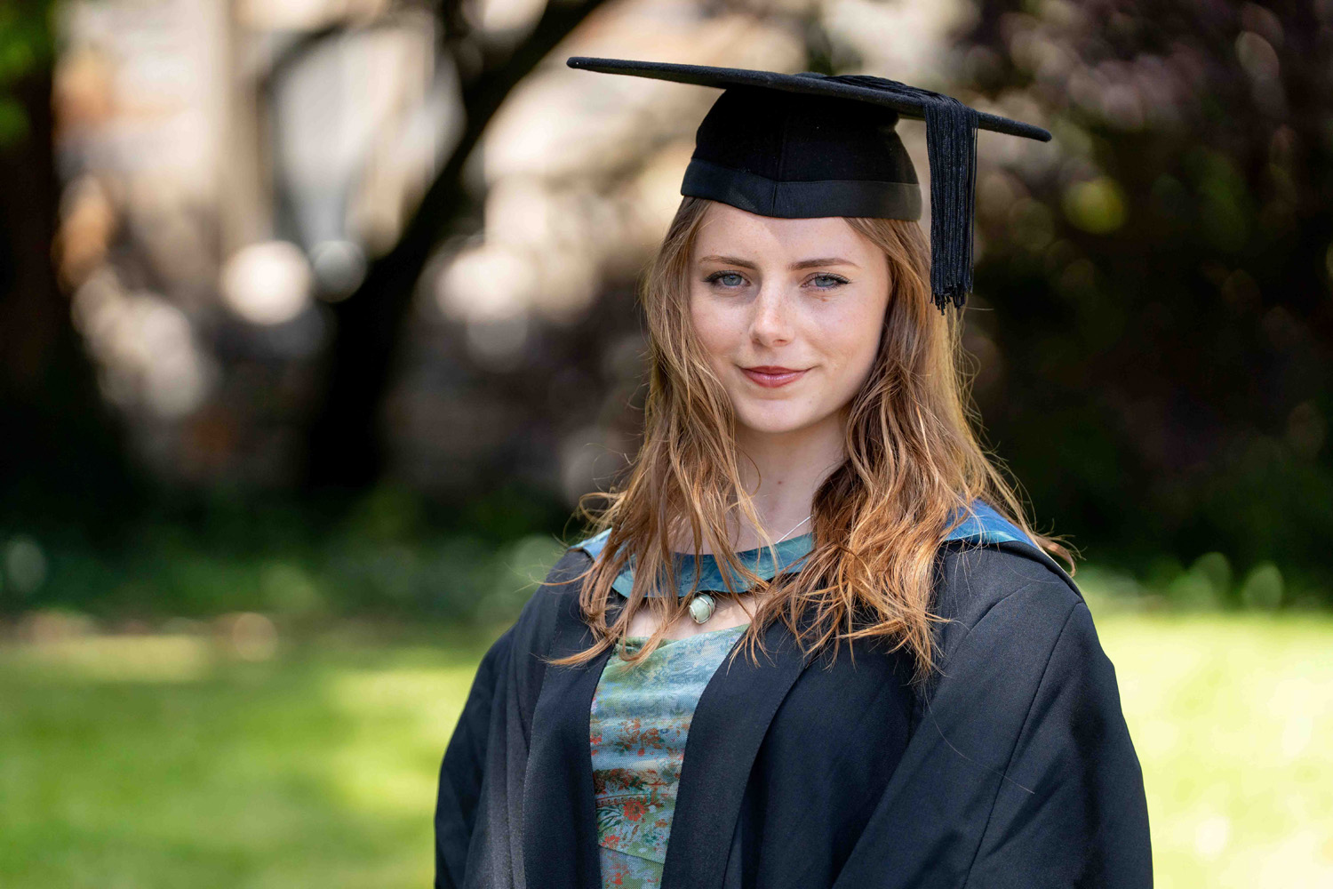 Young woman with long brown hair wearing a graduation cap and gown standing outdoors with a blurred natural background.