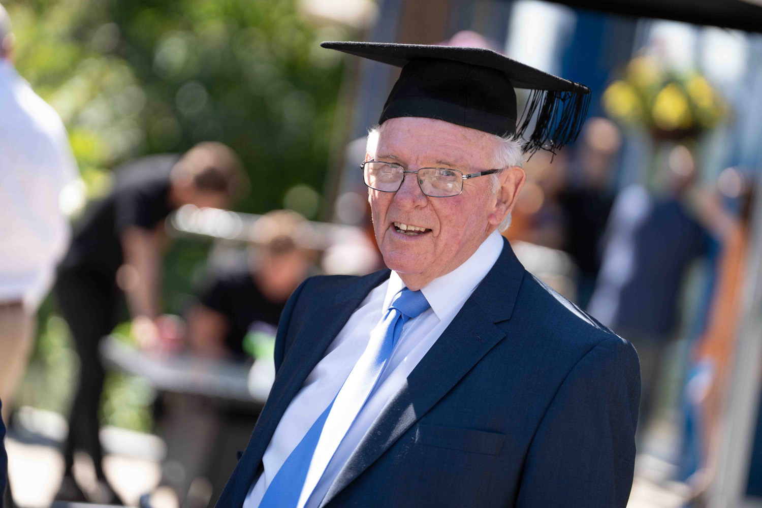 Elderly man wearing a dark suit with a light blue tie and a black graduation cap, smiling outdoors with blurred people and greenery in the background.