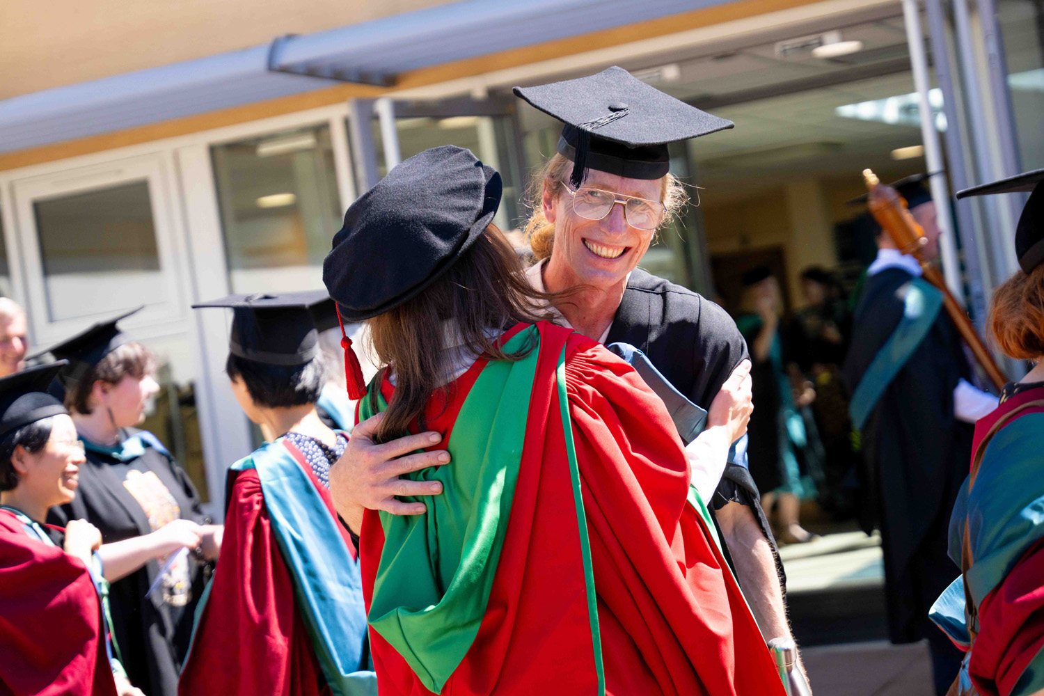 Two graduates in academic gowns and mortarboards embracing and smiling joyfully at an outdoor graduation ceremony with other graduates in the background.