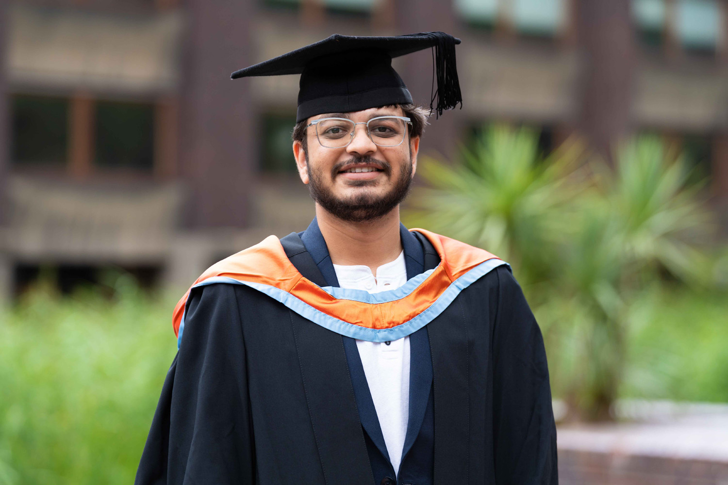 Young man wearing graduation cap and gown with an orange and blue hood, standing outdoors and smiling at the camera with blurred green plants and a building in the background