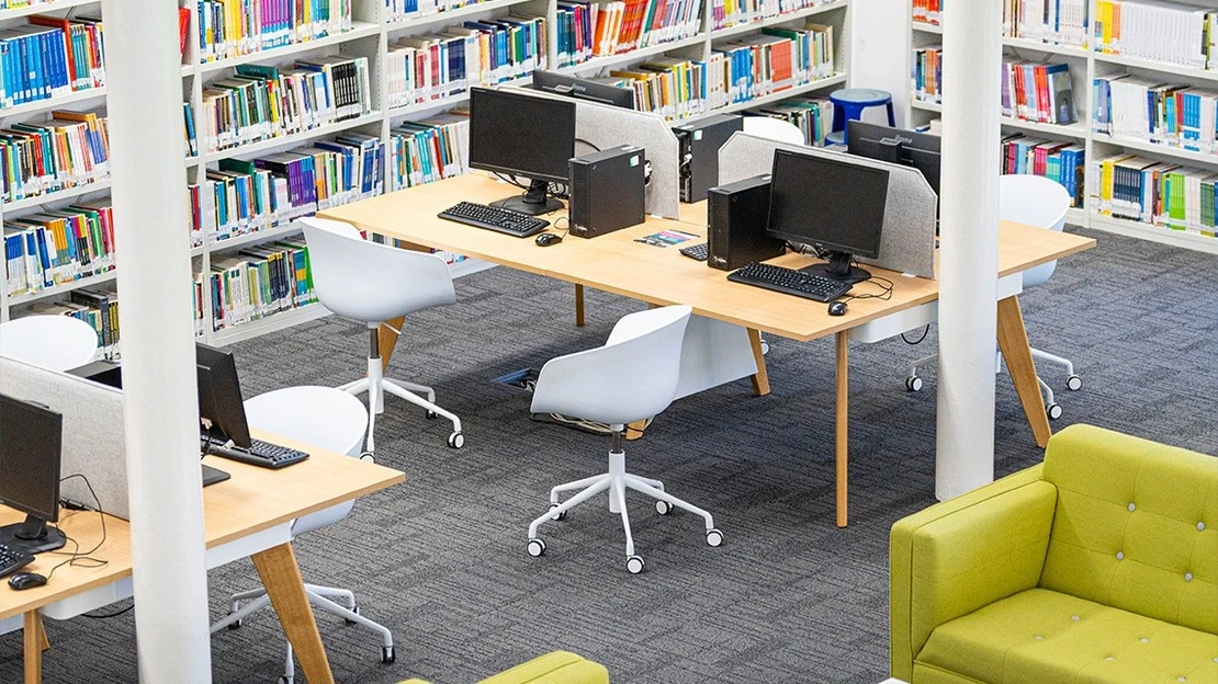 Modern library interior with wooden desks equipped with computers, white swivel chairs, gray carpet, and green upholstered couch, surrounded by bookshelves filled with colorful books.