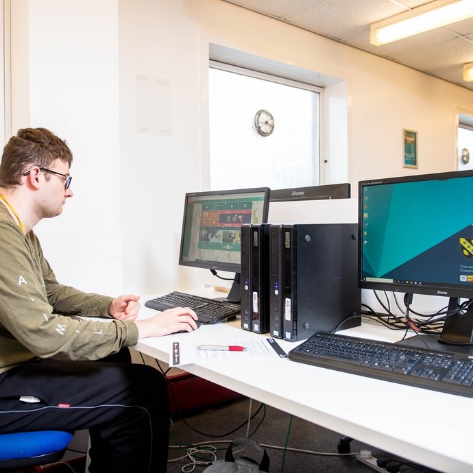 A man with glasses and a hearing aid sitting at a desk using a computer keyboard and mouse, with two desktop monitors and computer towers on the desk in a brightly lit office environment with large windows and a wall clock outside the window.