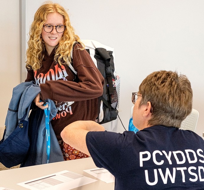 A young woman with curly blonde hair and glasses wearing a brown sweatshirt and carrying a backpack is smiling and interacting with a seated person wearing a navy blue shirt with white text at a desk with papers on it.