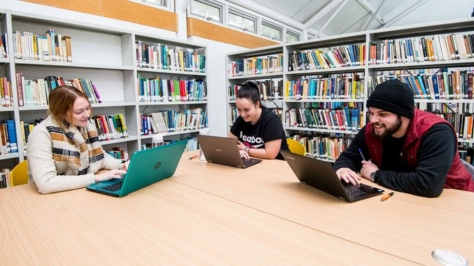 Three people sitting at a wooden table in a library with laptops, surrounded by shelves filled with books, two women and one man engaged in working and smiling.