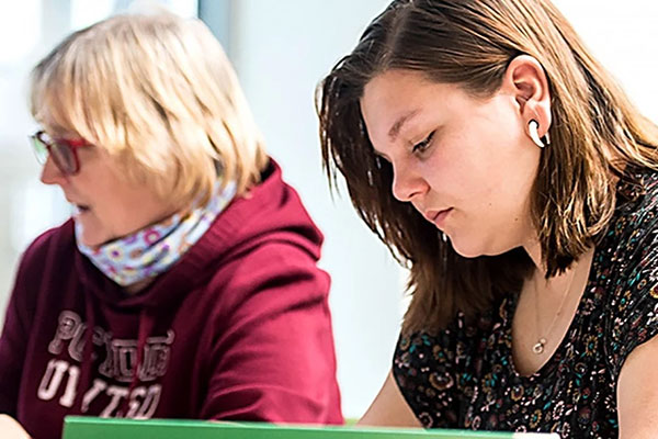 Two women sitting indoors, one with short blonde hair wearing glasses and a maroon hoodie looking to the side, and the other with shoulder-length brown hair wearing a patterned shirt and white wireless earbuds focused on an activity.