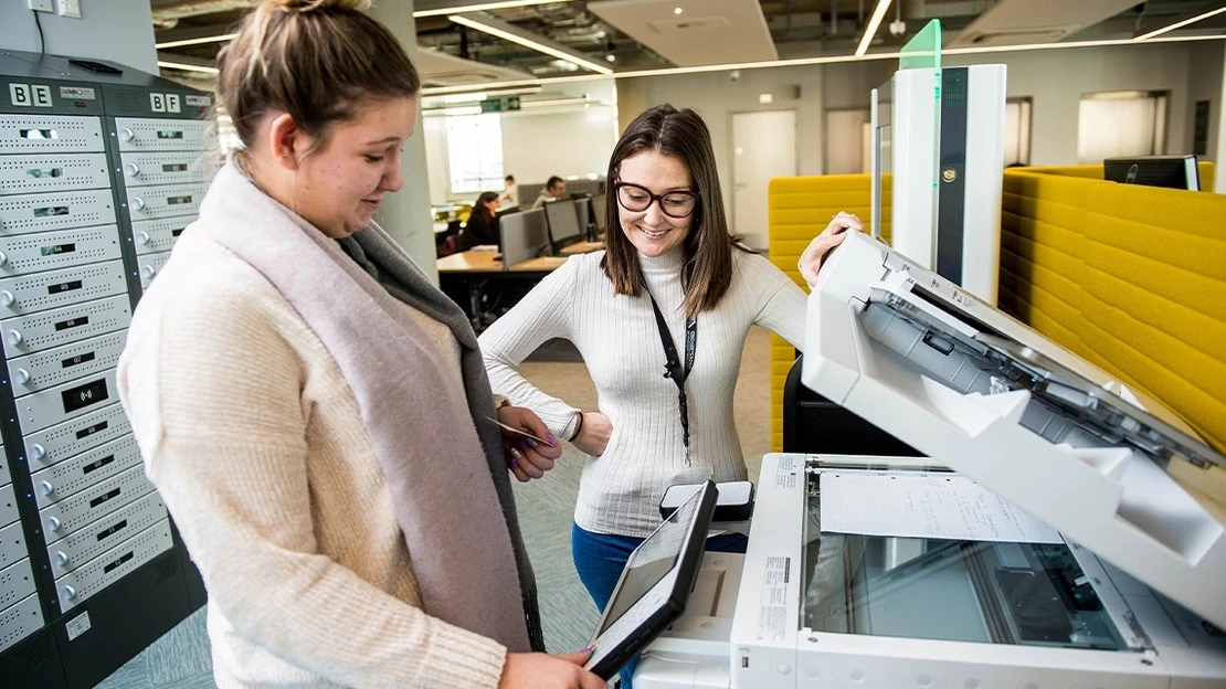 Two women using a photocopier in a modern office setting, one holding a tablet and the other lifting the copier's lid.