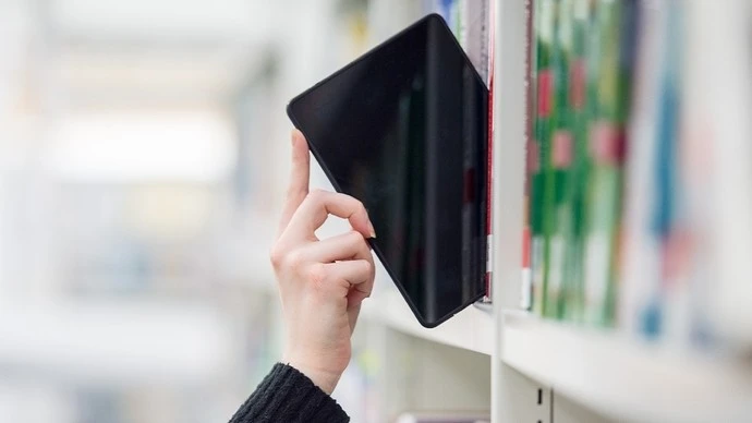 Hand removing or placing a black tablet device among books on a white bookshelf in a library or bookstore.