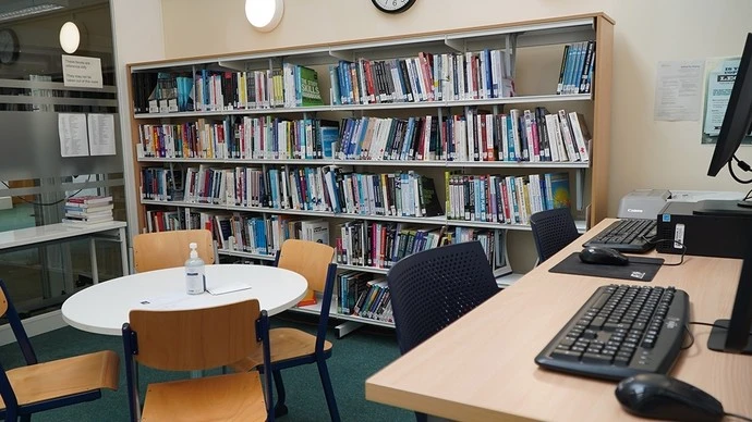 Interior of a library with shelves filled with books, a round table surrounded by wooden chairs, and a desk with computer keyboards, monitors, and a bottle of hand sanitizer on the table.