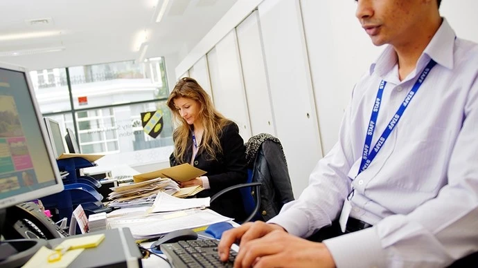 A male employee wearing a light purple shirt and staff lanyard typing on a keyboard in the foreground, with a female colleague sitting at a cluttered desk in the background reviewing documents inside a modern office.