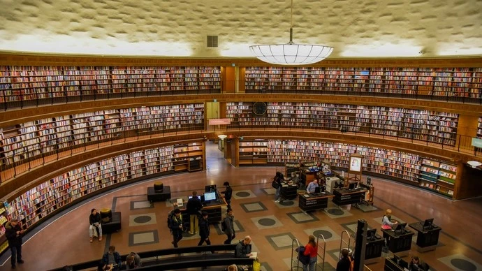 Interior of a large circular library with curved wooden bookshelves filled with books on two levels, people walking and sitting in the central open space, and a large circular light fixture hanging from the ceiling.