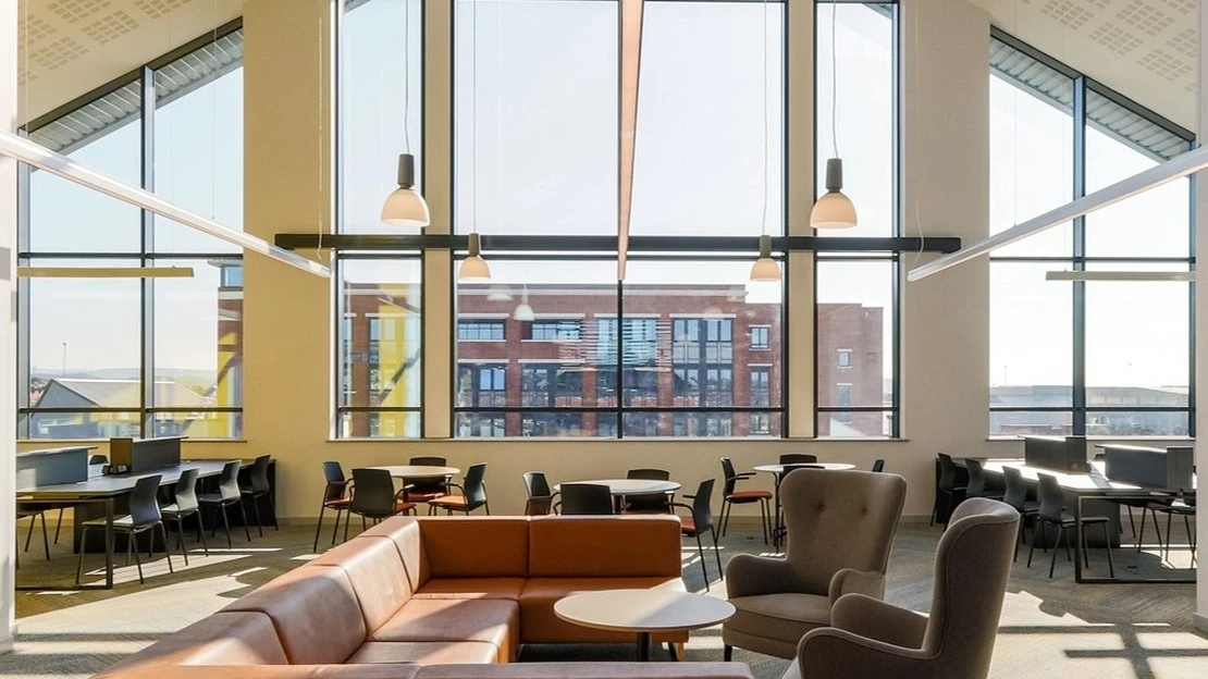 Modern office lounge area with large floor-to-ceiling windows, brown leather sectional sofa, gray armchairs, round tables, and rows of black chairs at long desks under hanging pendant lights.