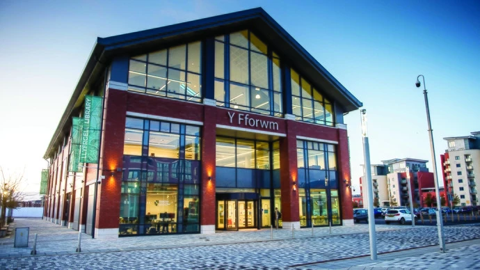Modern library building named Y Fforwm with large glass windows, red brick facade, illuminated from inside at dusk, situated on a paved street with parked cars and nearby apartment buildings