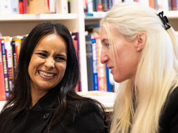 Two women sitting and interacting indoors with bookshelves filled with books in the background; the woman on the left with dark hair is smiling broadly, while the woman on the right with long blonde hair looks towards her.