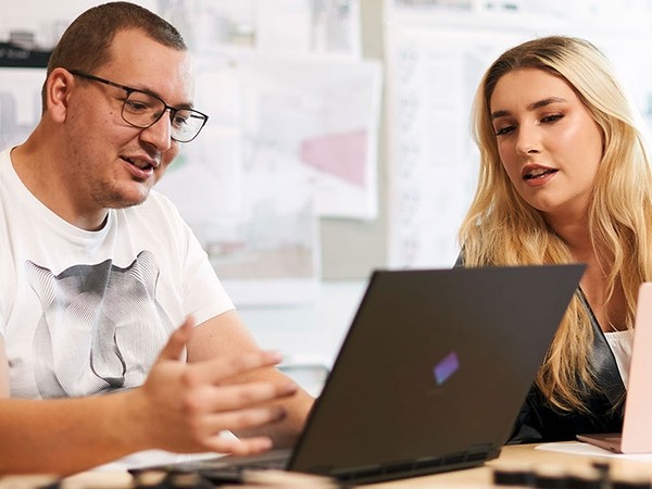 Two people, a man with glasses and a woman with long blonde hair, sitting and looking at a laptop screen together in an office setting.