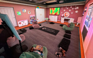 A cozy classroom with pink walls, bean bag chairs with stuffed animals, a TV, and a large rug with "University of Wales" text.